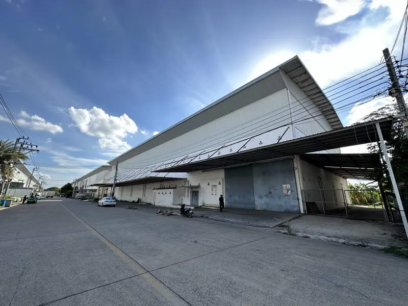 Large industrial warehouse with a wide entrance and corrugated metal roof under a blue sky. Power lines run beside the street.