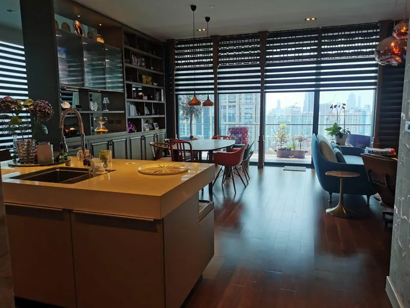Open-concept kitchen and living area with modern furniture, a large window overlooking a cityscape, and a dining table under pendant lights.