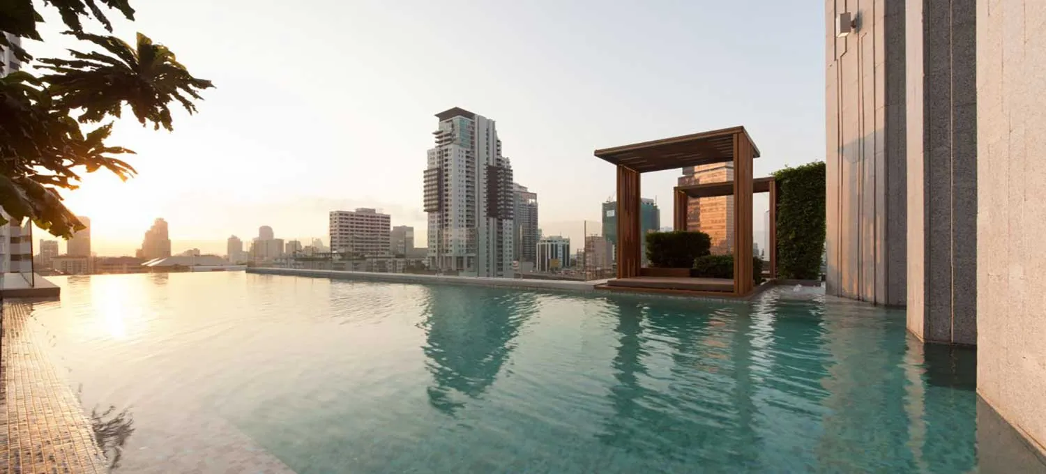 Rooftop infinity pool overlooking a city skyline at sunset, with modern skyscrapers and a wooden cabana.