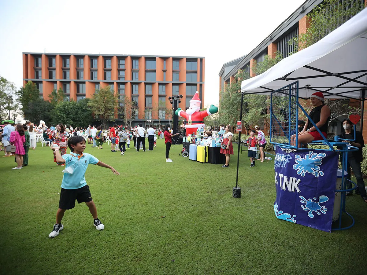 A child throws a ball at a dunk tank while people gather on a grassy area with a large Santa Claus inflatable decoration.