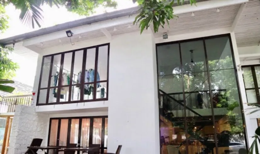 Modern two-story building with large windows, surrounded by trees. Bottom level has a dining table set under a wooden ceiling, and stairs are visible through a tall glass panel.