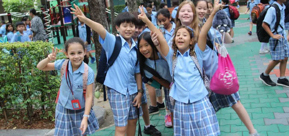 Group of smiling children in school uniforms posing playfully outdoors, with backpacks and trees in the background.