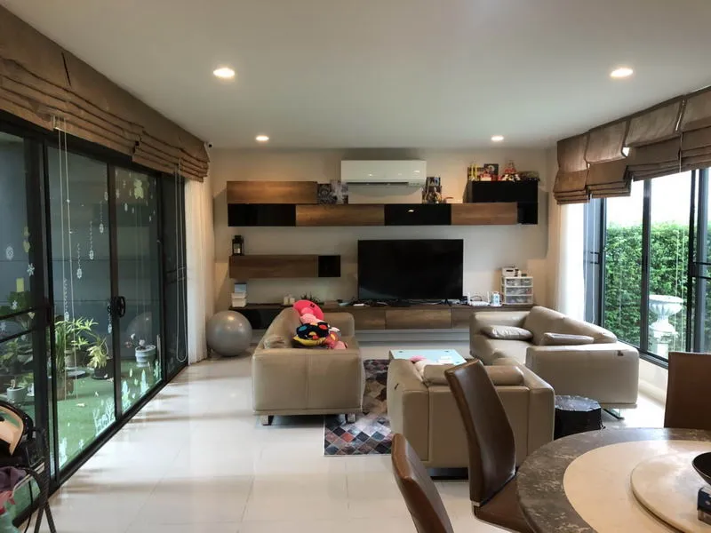 Modern living room with beige sofas, a TV, wooden shelves, and large windows overlooking a garden.