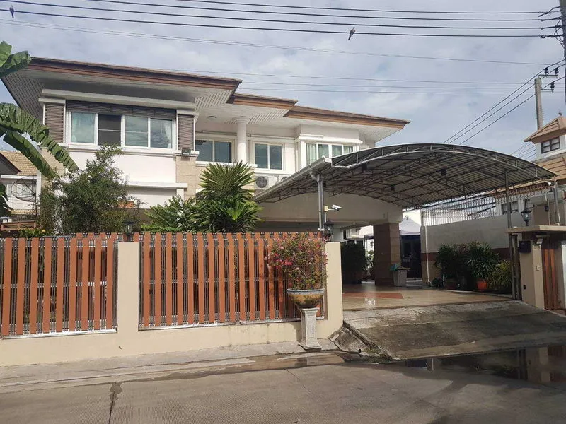 Two-story house with a gated driveway, metal carport, and lush plants behind a wooden fence.