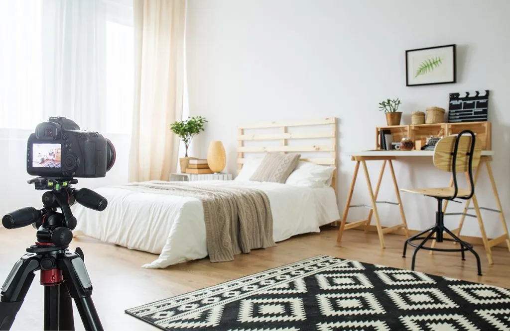 Camera on a tripod in a bright bedroom with a bed, desk, and patterned rug.