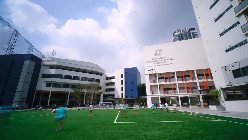 Modern school campus with students playing on a green field, surrounded by white and blue buildings under a partly cloudy sky.
