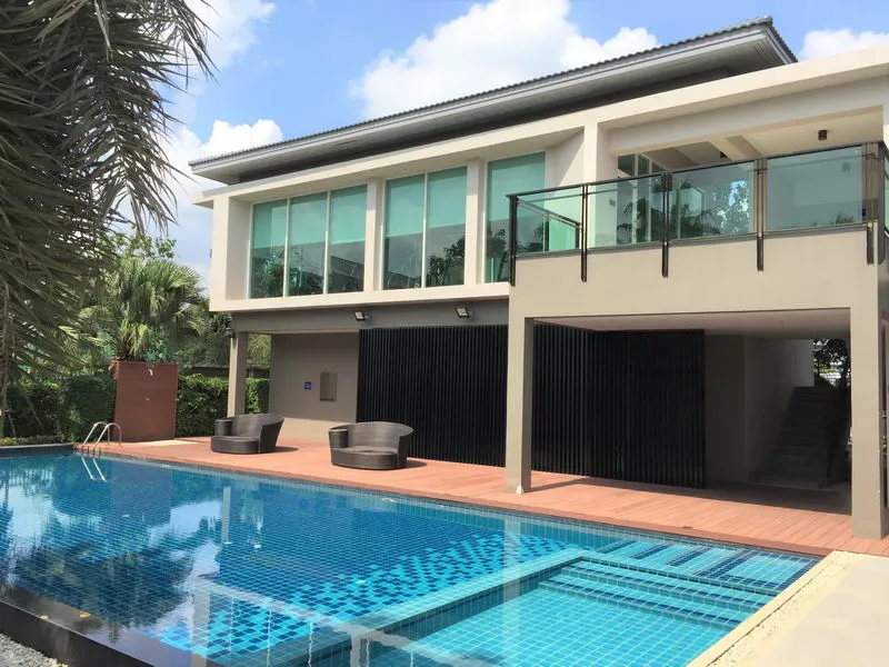 บ้านสไตล์โมเดิร์น มีหน้าต่างบานใหญ่และระเบียง มองเห็นสระว่ายน้ำที่ล้อมรอบด้วยพื้นไม้