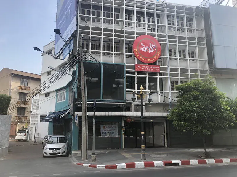 Street view of a multi-story building with a red sign featuring a dragon logo. There's a small car parked outside and trees lining the sidewalk.