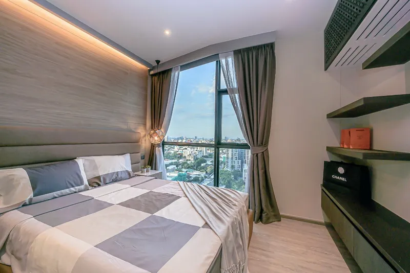 Modern bedroom with a large bed, quilt, and pillows next to a window with cityscape views. The room includes shelves with decorative items.
