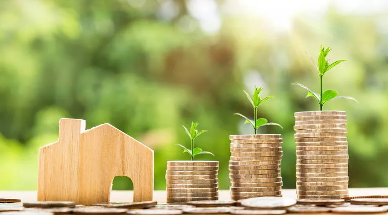 Wooden house model and stacked coins with growing plants on top, symbolizing investment growth.