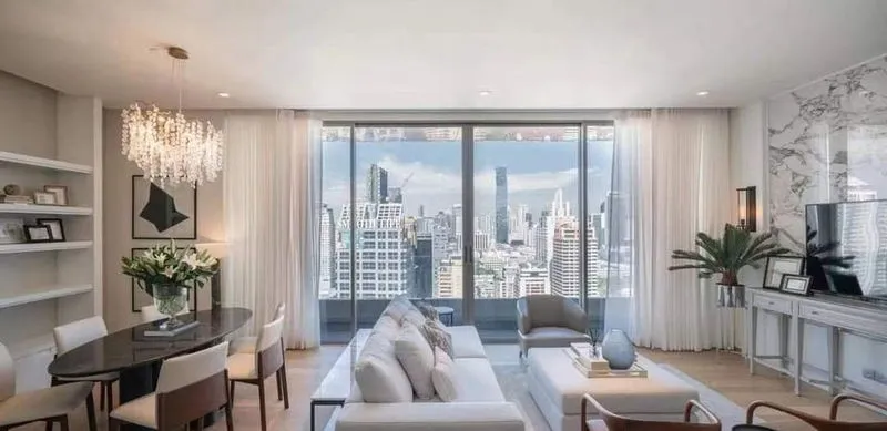 Modern living room with a large window view of a city skyline, featuring a chandelier, sectional sofa, dining table, and wall-mounted TV.