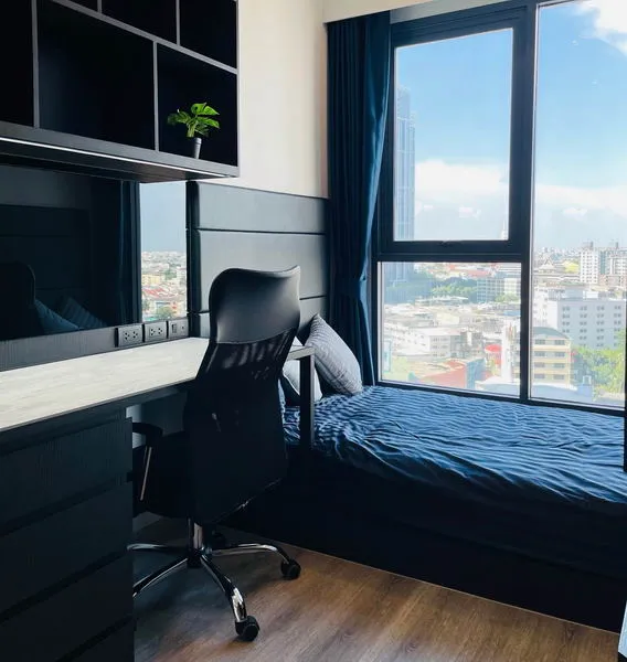 Compact modern room with a single bed, desk, office chair, shelves, and large window offering a city view.