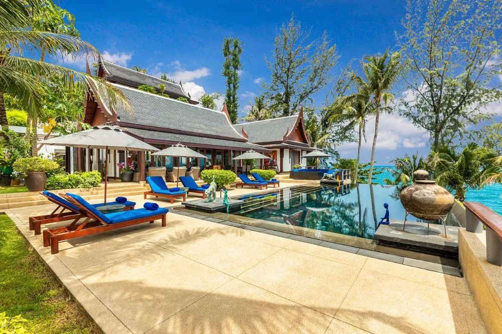 Luxury tropical villa with a modern infinity pool, surrounded by palm trees and overlooking the ocean, featuring blue sun loungers and sun umbrellas.