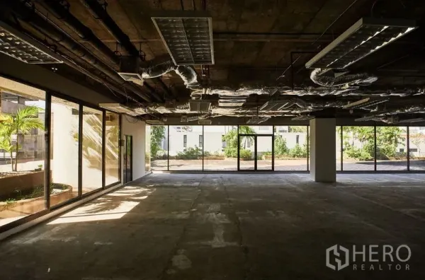 Office Space for rent in Bangkok - Open office area with floor-to-ceiling glass facing a landscaped terrace, exposed ceiling and concrete floor.