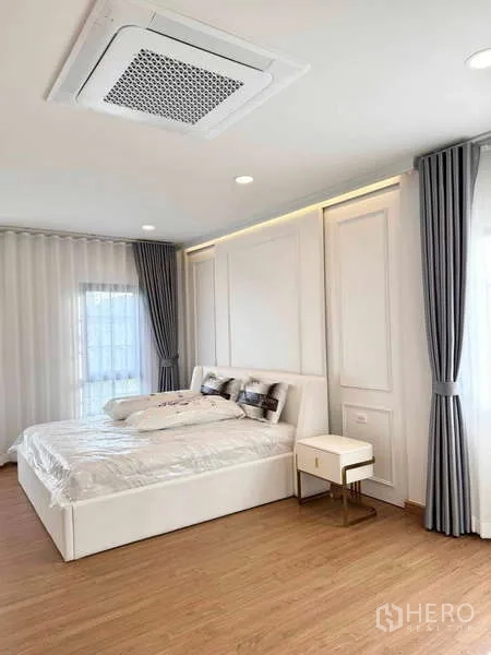 Detached House for rent or sale in Bang Phli, Samut Prakan - Master bedroom with panel accent wall, white bed, cassette ceiling AC, wood flooring and corner windows.