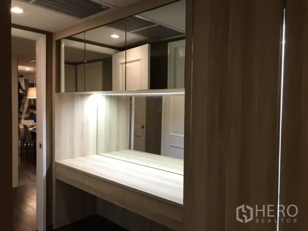 Condominium for rent or sale in Pathum Wan, Bangkok - Bright dressing nook with illuminated mirror, upper cabinets, and wood cabinetry along the corridor.