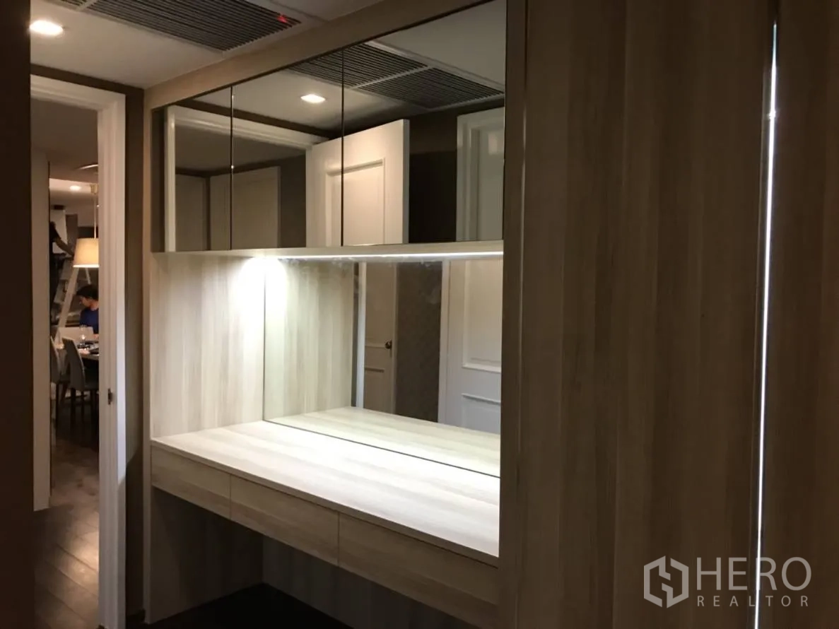 Condominium for rent or sale in Pathum Wan, Bangkok - Bright dressing nook with illuminated mirror, upper cabinets, and wood cabinetry along the corridor.