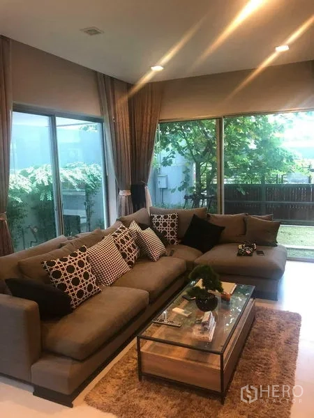 Detached House for rent or sale in Bang Kapi, Bangkok - L-shaped sofa in the living room facing a glass coffee table and garden view.