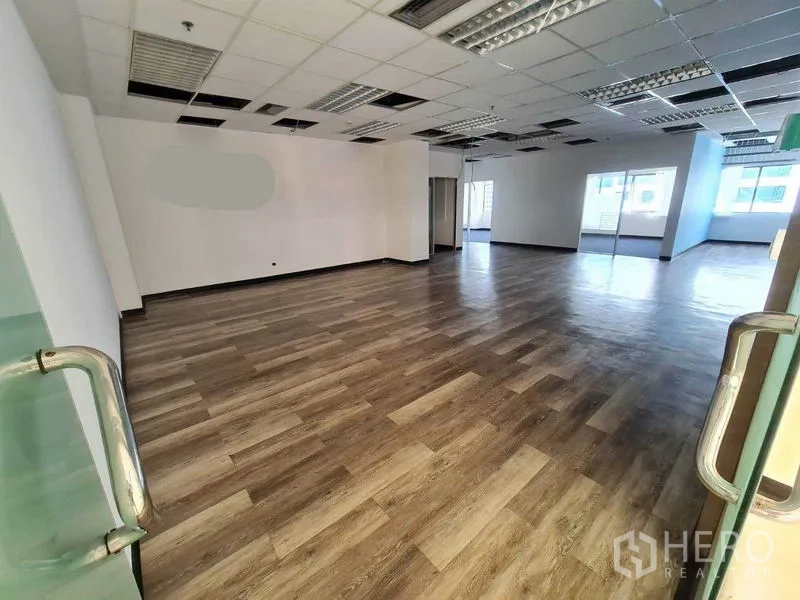 Office Space for rent in Watthana, Bangkok - Open-plan office interior with wood-look flooring, grid ceiling lights and large windows, viewed from glass doors.