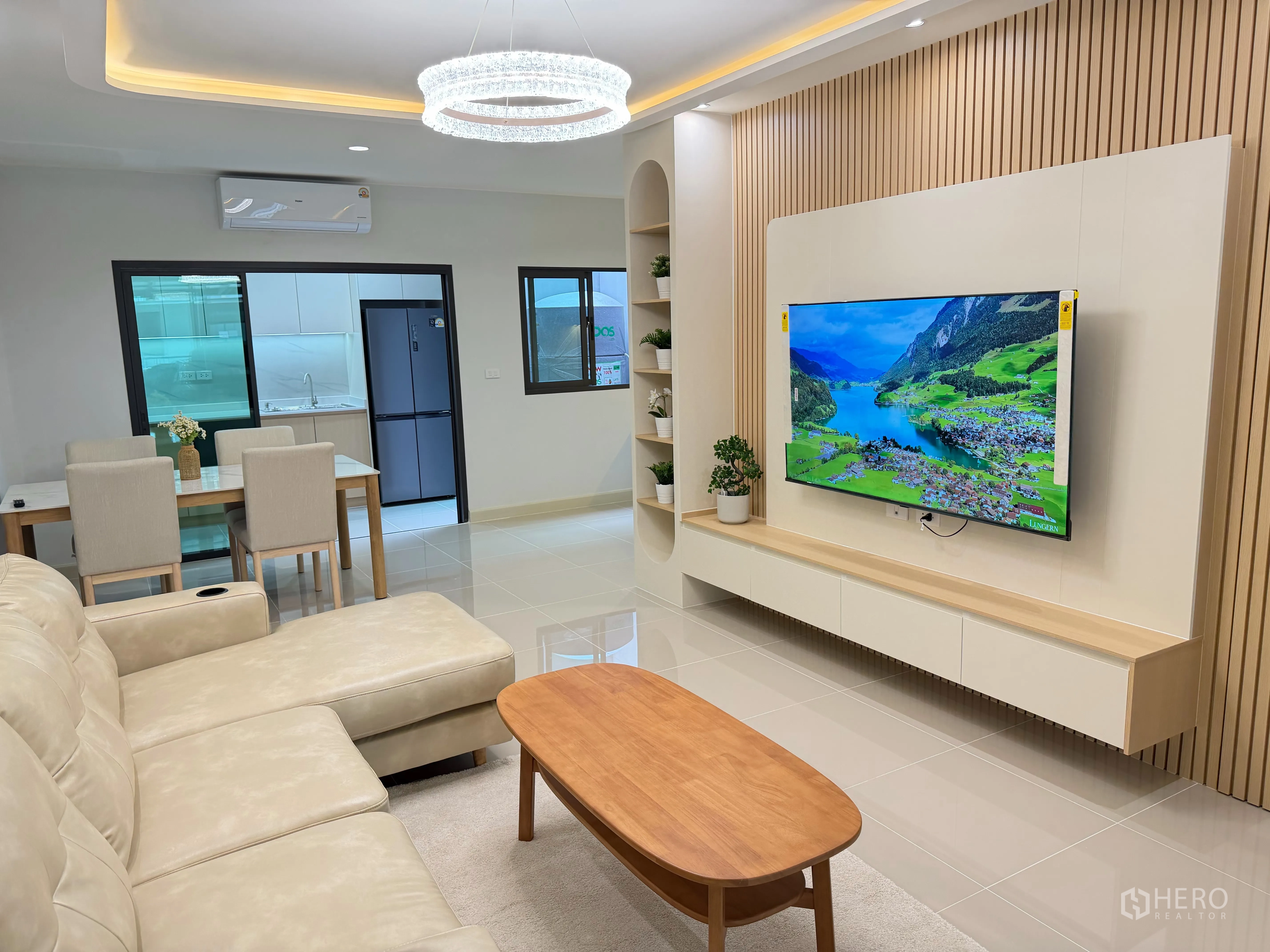 Townhouse for rent in Bang Phli, Samut Prakan - Bright open-plan living room with L-shaped sofa, wall-mounted TV on a wood accent wall, dining set, and a separate kitchen with refrigerator.