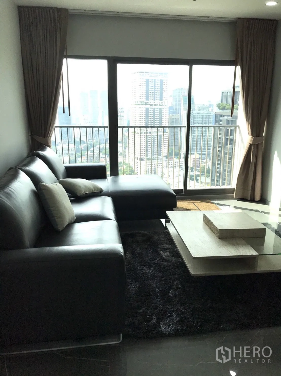 Condominium for rent in Khlong Toei, Bangkok - Living area with black L-shaped sofa facing a balcony and city skyline views.