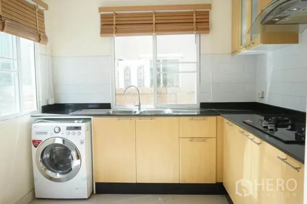 Detached House for rent in Bang Phli, Samut Prakan - Modern kitchen with light wood cabinetry, black countertops, gas hob, sink and washing machine under the window.