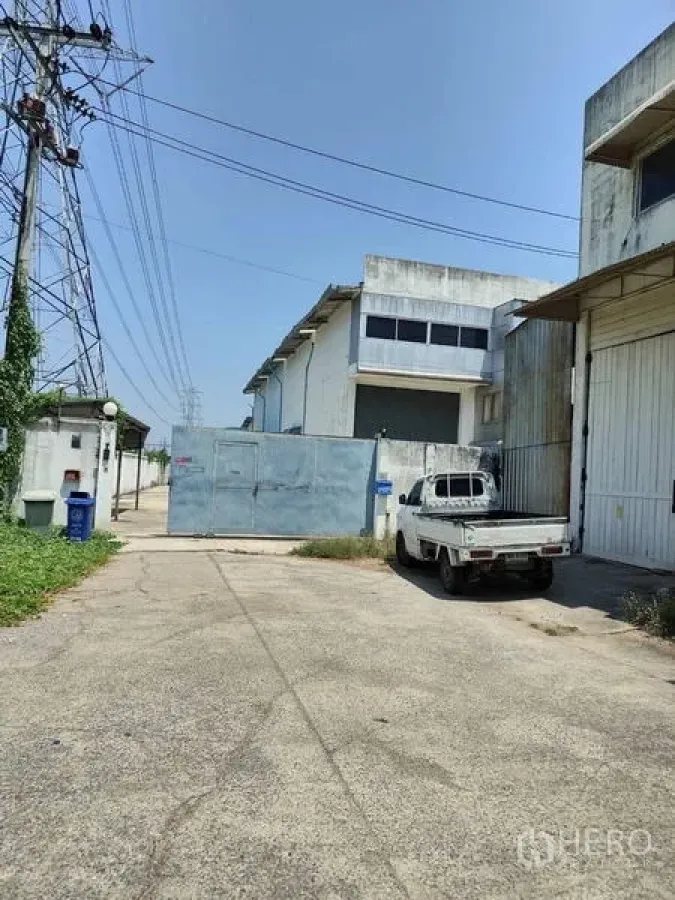 工厂 出售在Sai Noi, Nonthaburi - 工厂入口，金属大门、白色皮卡与宽阔混凝土车道，旁有高压线。