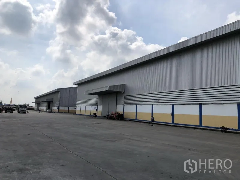 Warehouse for rent in Phra Nakhon Si Ayutthaya - Wide concrete yard facing long steel-clad warehouse blocks under a cloudy sky in Bang Sai.