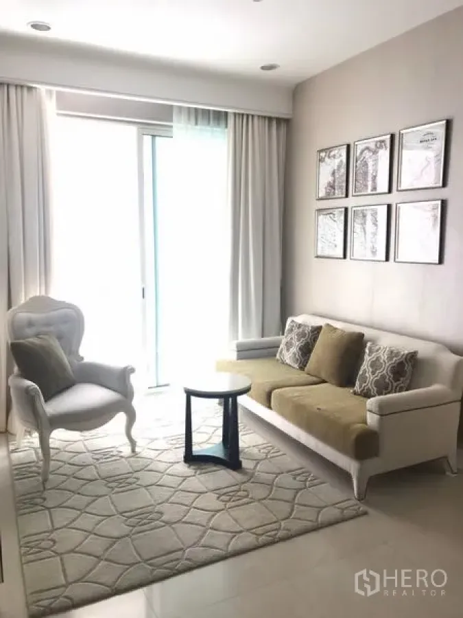 Condominium for rent in Pathum Wan, Bangkok - Bright condo living room with white sofa, accent armchair, patterned rug and sliding glass door with sheer curtains.