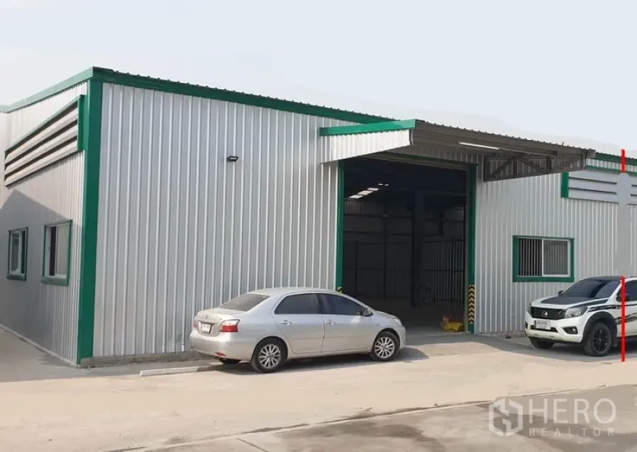 Warehouse for rent in Pak Kret, Nonthaburi - Metal-clad warehouse exterior with green trim, wide roller shutter, and parking area with cars.