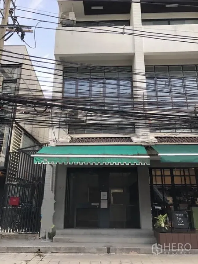 Office Space for rent in Watthana, Bangkok - Three-story shophouse-style office building facade with green awning, glass doors, metal window grilles, and overhead cables on an urban street.