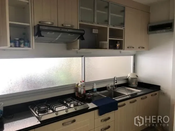 Townhouse for rent or sale in Suan Luang, Bangkok - Suan Luang townhouse kitchen with built-in light-wood cabinets, gas hob, range hood and long black countertop under frosted windows.