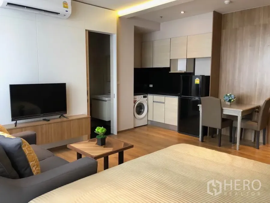 Condominium for rent in Khlong Toei, Bangkok - Open-plan living area with kitchenette, washing machine, and TV on wood flooring.