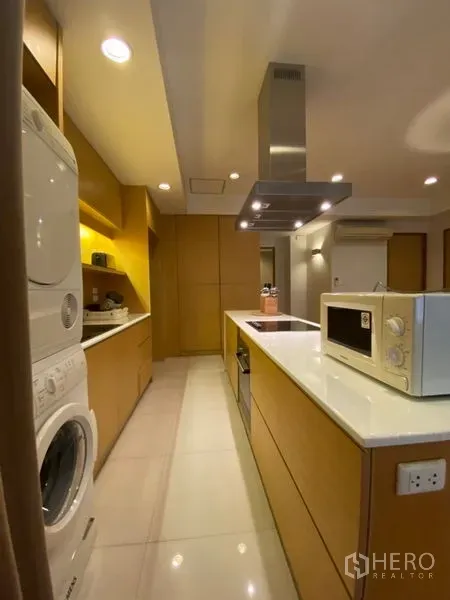 Condominium for rent in Watthana, Bangkok - Galley kitchen featuring a white island with cooktop and hood, built-in cabinets, and stacked washer-dryer.