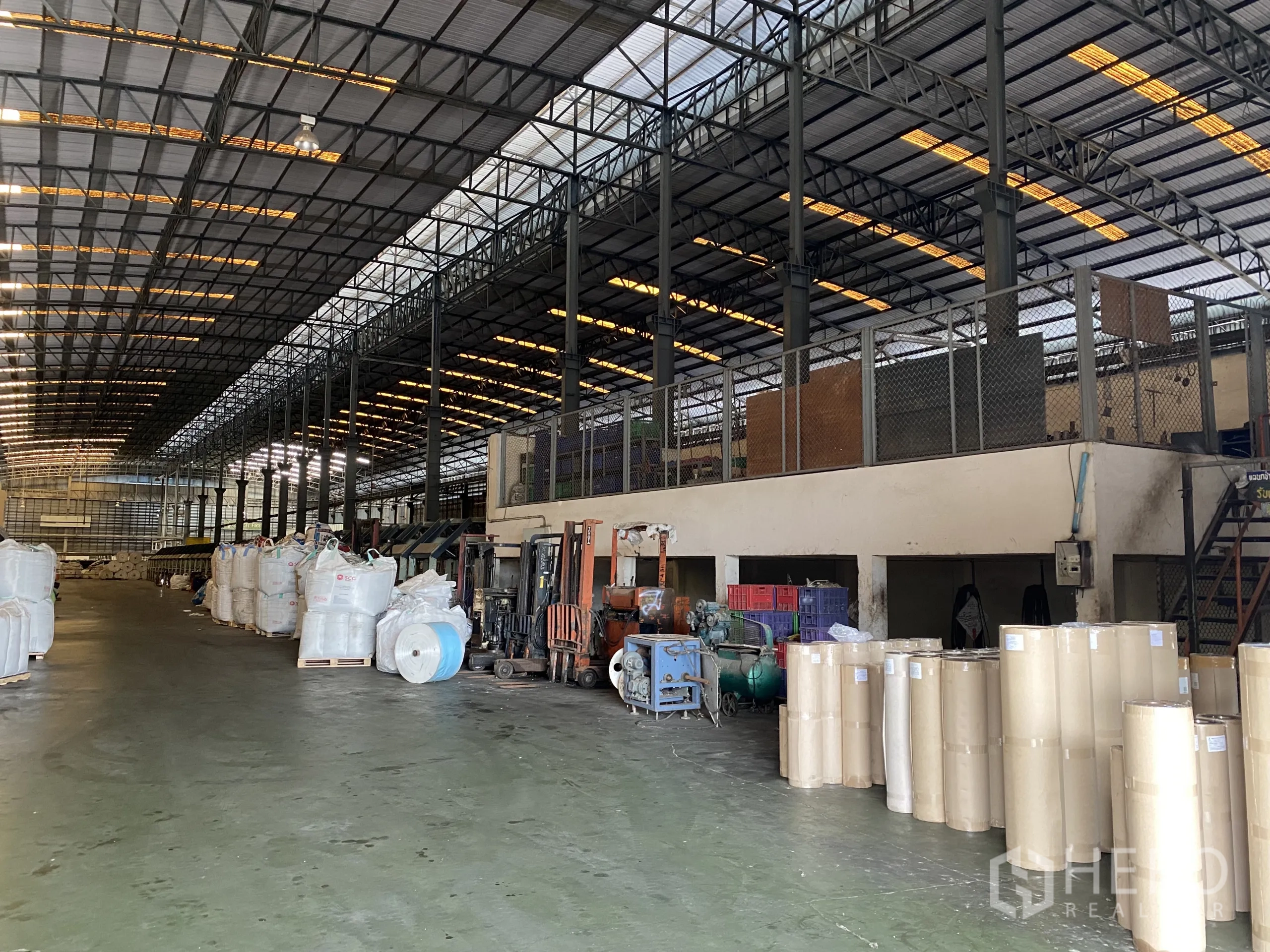 Factory for rent in Ban Phaeo, Samut Sakhon - Warehouse interior with forklifts, mezzanine office and stacked industrial materials under a steel roof.