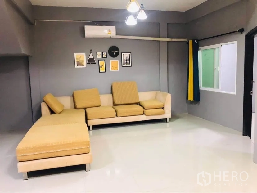 Townhouse for rent or sale in Huai Khwang, Bangkok - Spacious living room with L-shaped sofa, grey feature walls, air conditioner and wall art.