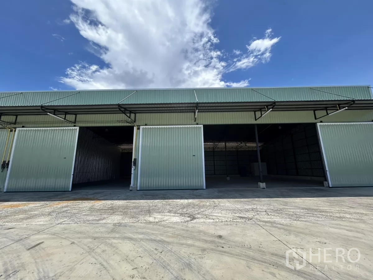 Warehouse for rent in Ban Pho, Chachoengsao - Row of green metal warehouses with wide sliding doors, covered loading canopies and a large concrete yard under blue sky.
