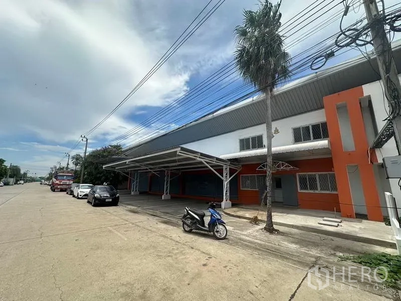 โรงงาน ให้เช่าในNonthaburi - ภาพมุมเฉียงของอาคารสีส้มขาว กันสาดโลหะ และถนนหน้าอาคารกว้าง.