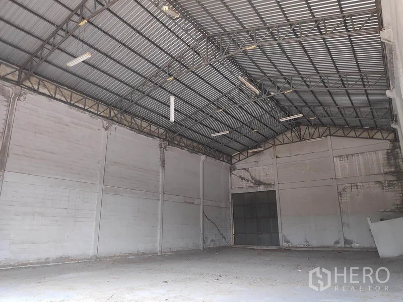 Factory for rent in Sai Noi, Nonthaburi - Empty high-ceiling factory interior with steel truss roof, concrete floor and a large sliding door.