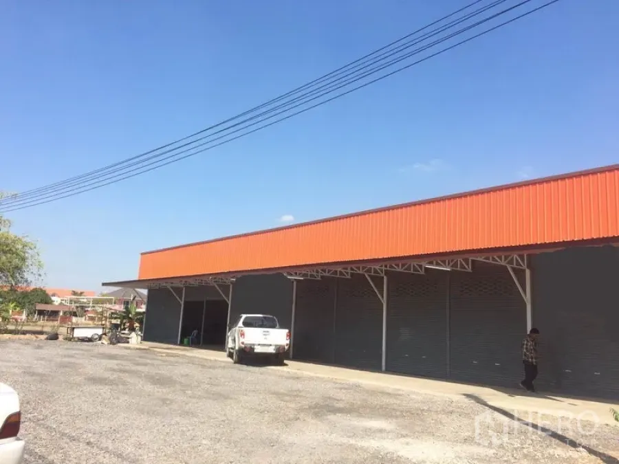 Warehouse for rent in Pathum Thani - Warehouse exterior with orange fascia, multiple roller shutter doors, and a wide gravel parking and loading area in Lam Luk Ka.