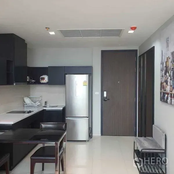 Condominium for rent in Khlong Toei, Bangkok - Modern open kitchen with black cabinetry, dining set and a stainless-steel refrigerator by the entrance.