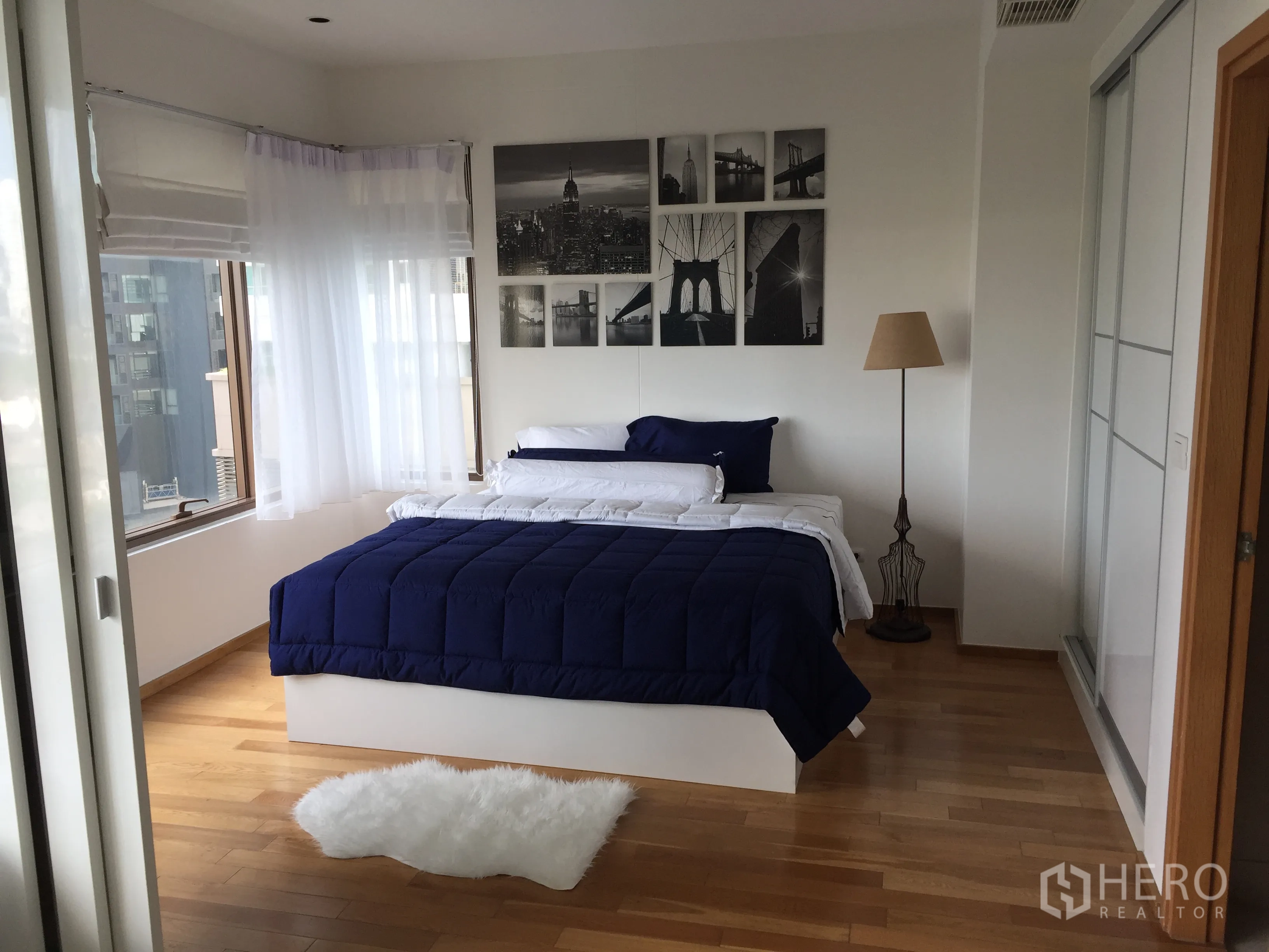 Condominium for rent in Khlong Toei, Bangkok - Sunlit bedroom with corner windows, blue duvet, sheer curtains, wood flooring and wall art above the bed.