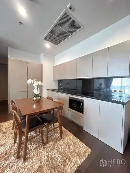 Condominium for rent or sale in Watthana, Bangkok - Modern kitchen with black stone backsplash, built-in appliances and a wooden dining table on a shag rug.