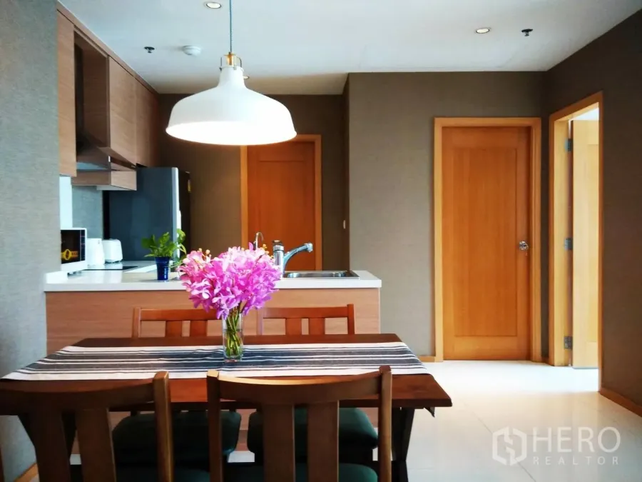 Condominium for rent or sale in Khlong Toei, Bangkok - Open-plan kitchen and dining area with pendant light, wooden cabinetry, countertop sink and a four-seat table with flowers.