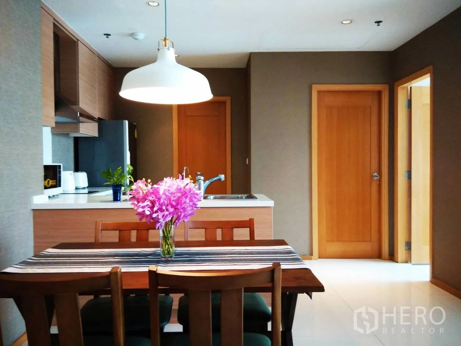 Condominium for rent or sale in Khlong Toei, Bangkok - Open-plan kitchen and dining area with pendant light, wooden cabinetry, countertop sink and a four-seat table with flowers.