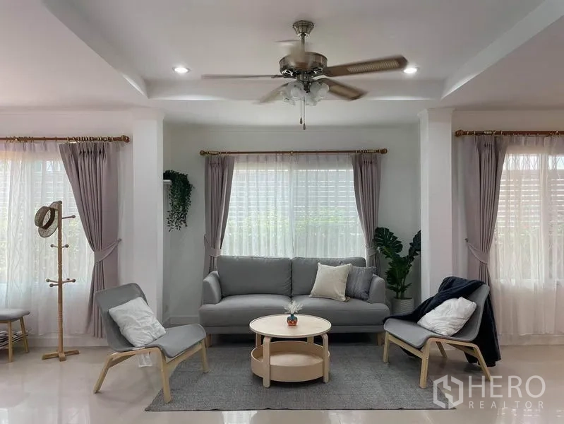 Detached House for rent in Suan Luang, Bangkok - Bright living room with ceiling fan, gray sofa set, armchairs and large curtained windows.
