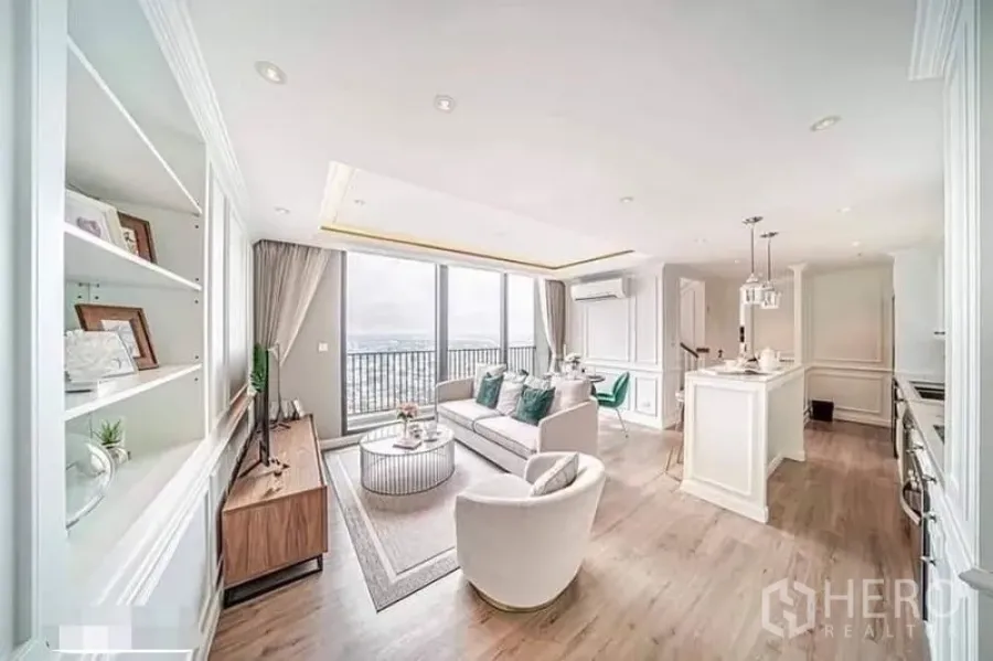 Condominium for rent or sale in Watthana, Bangkok - Sunlit living room with built-in shelving, sofa set and skyline view from tall glass walls.