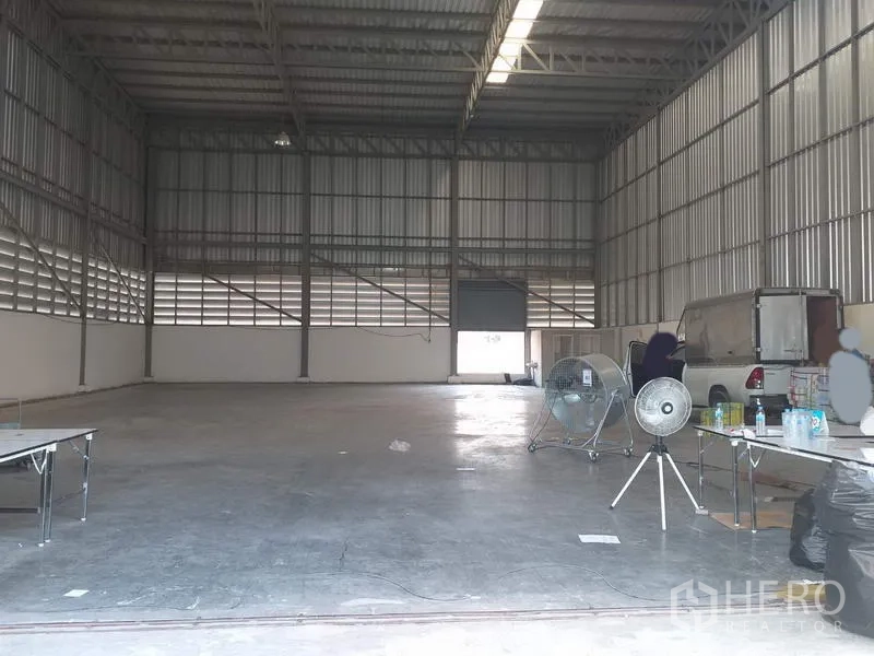 Warehouse for rent in Pak Kret, Nonthaburi - Spacious warehouse interior with a high steel roof, skylight strip, roller shutter, and smooth concrete floor.