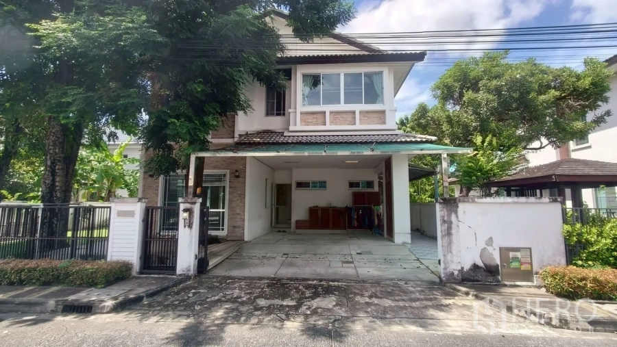 Detached House for rent or sale in Mueang Samut Prakan, Samut Prakan - Two-storey detached house facade with gated entrance and covered carport shaded by trees.