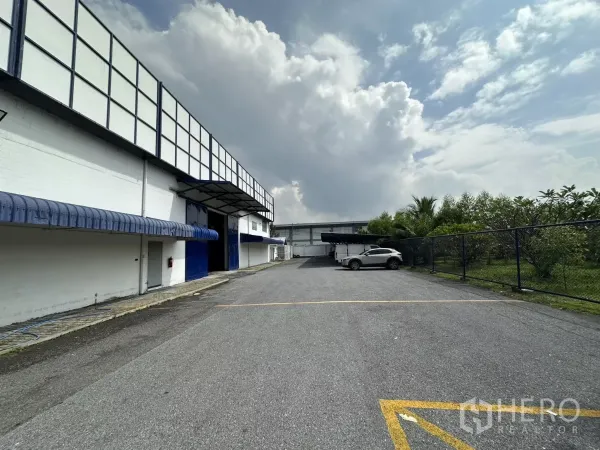 Warehouse for rent in Phra Khanong, Bangkok - Spacious driveway and on-site parking beside the warehouse with fence and greenery, showing loading bay access.